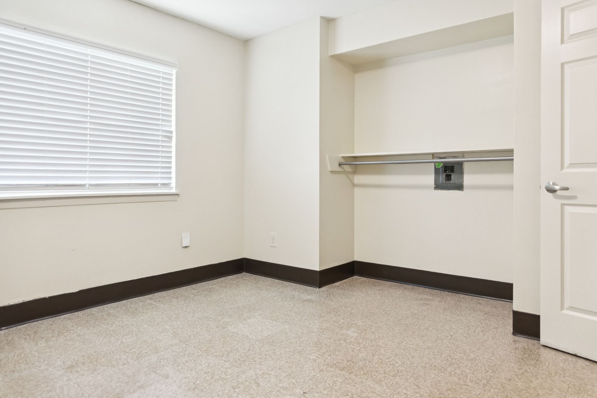 A room with a white door, a window with blinds, and a shelf with a black box on it.